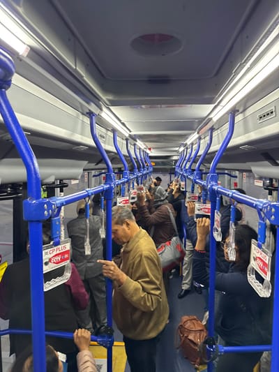 The inside of a bus is shown, with passengers using blue handrails. Many people are engrossed in their phones. Signage labeled "CP PLUS" is attached to the handrails. The bus interior is brightly lit with long, horizontal lights on the ceiling. A door with a circular window is visible on the left side. The overall scene depicts a typical commute on a crowded bus.