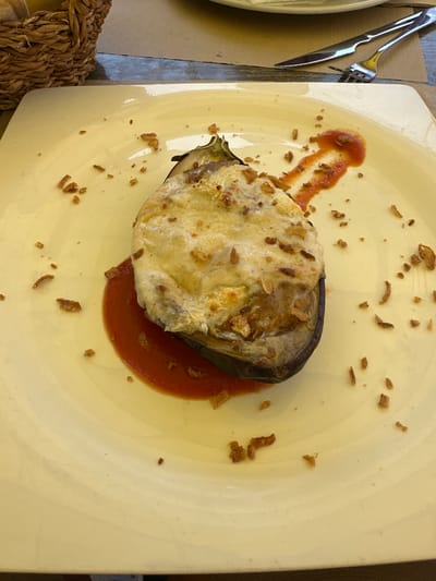 A top-down, medium shot captures a single serving of baked eggplant on a large, pale yellow plate. The eggplant is halved, with its flesh scooped out and then re-filled and baked, topped with a golden-brown melted cheese crust. A vibrant red sauce pools beneath and around the eggplant. Scattered across the plate are small, dark brown crispy bits, possibly fried onions or herbs. To the left of the plate, a fork and knife are laid out, their metallic sheen catching the light. The background hints at a table setting with a woven placemat visible in the lower left corner and a glimpse of a textured brown surface on the right. The lighting suggests daytime, and the overall scene conveys a simple, rustic meal presentation. No visible text is present. The location context provided is Santa Cristina d'Aro, Spain.