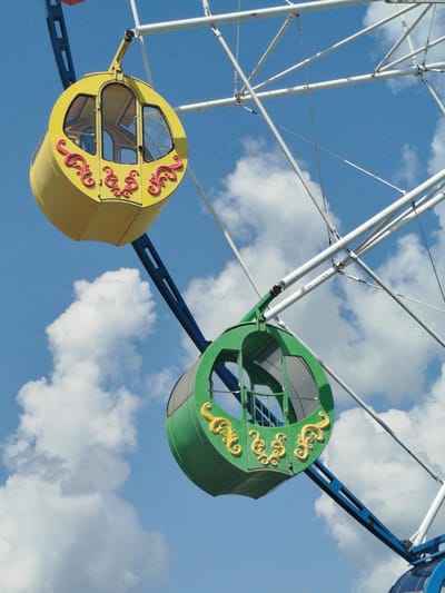 The image shows two gondolas of a Ferris wheel against a backdrop of blue sky and white clouds. The gondolas are suspended from the white metal structure of the Ferris wheel. One gondola is yellow with red decorative elements, and the other is green with gold decorative elements. The Ferris wheel is likely located in Yalutorovsk, Russia, as it is a common amusement park attraction. There are no people visible in the image. The lighting suggests it is daytime, possibly mid-morning or afternoon.