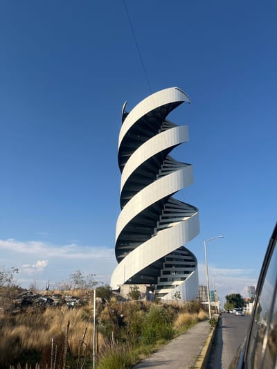 The image shows a modern, white, spiral-shaped building complex against a clear blue sky in Tlaxcalancingo, Mexico. The building's design resembles a twisting ribbon or DNA helix. Adjacent to the building is a landscape of dry vegetation, rocks, and scattered debris. A road with street lights and distant buildings is visible in the background. The perspective suggests the photo was taken from a moving vehicle.
