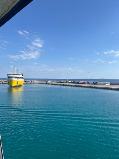 A bright yellow and white ferry is docked at the port of Kyllini, Greece. The ferry is positioned on the left side of the image, facing the viewer. The water in front of the ferry is a vibrant turquoise with small ripples. A concrete pier extends from the right side of the image, lined with various shipping containers and trucks. Several lampposts are also visible along the pier. In the distance, the horizon is marked by a thin line of water separating the sea from a hazy mountainous landmass. The sky above is a clear blue with scattered white clouds. The overall scene depicts a bustling port on a sunny day.