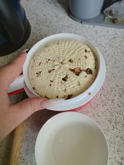 A person's hand with long, light-colored artificial nails is holding a round, white cheese with a patterned surface and visible pieces of nuts embedded within it. The cheese is contained within a red and white mold or container. A white bowl is positioned in the foreground, partially obscuring the lower portion of the mold. The background shows a speckled countertop, a dark blue appliance, and a light gray object. The scene appears to be a kitchen environment. The image does not contain any information or visual cues that would allow for the identification of its location in Pervouralsk, Russia.