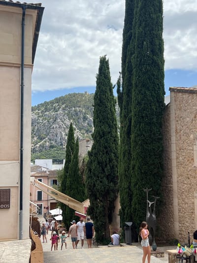 This image depicts a street scene in the town of Pollença, Spain, likely during daytime with a cloudy sky. In the foreground, a paved pathway slopes downwards, with people walking and standing. To the left, a light-colored building with a dark downspout dominates the frame. Further down the street, a row of tall, slender cypress trees lines the right side of the pathway. Beyond the trees, more buildings with terracotta roofs are visible, and in the background, a lush, green mountainside rises, topped with what appears to be a small castle or fortress. Several people are visible in the mid-ground, including families with children walking up the stairs and individuals browsing shops. A dark metal sculpture, abstract and resembling a stylized animal, stands near the cypress trees. A young woman in a pink top and denim shorts stands on the steps looking towards the sculpture. The overall atmosphere suggests a tourist destination, with shops and leisure activities.