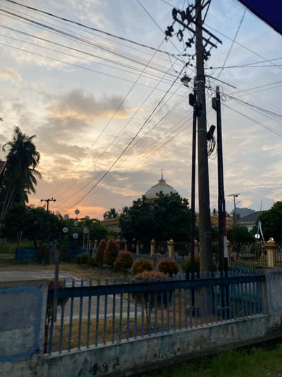 The image depicts a street scene at sunset in Uleejalan, Indonesia. In the foreground, a blue metal fence with a concrete base runs horizontally across the frame. Behind the fence, a patch of dry grass is visible. The middle ground features a complex network of power lines and utility poles, with one particularly large pole dominating the right side of the image. Several smaller decorative lights are also visible on poles further back.

Beyond the utility poles, a building with a prominent dome, likely a mosque, is situated. The dome is white with yellow accents and a small finial on top. The building appears to be surrounded by trees and foliage. A few palm trees are visible on the left side of the frame. The sky is filled with clouds, tinged with orange and yellow from the setting sun, creating a dramatic sunset backdrop. A person is briefly visible in the background near the mosque. The overall impression is a somewhat cluttered urban environment under a colorful sky.