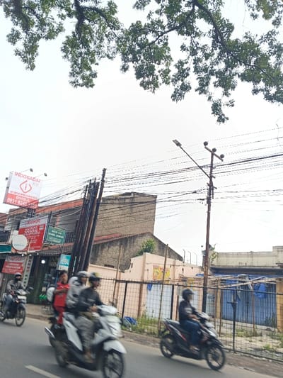 A street scene showcases several motorcycles in motion, with riders wearing helmets. A power pole laden with wires stands to the right. Buildings line the background, including one with a large "INDOGADAI" sign and smaller advertisements. Trees overhang from above, partially obscuring the sky. A fence encloses a structure with blue tarps, adding to the urban atmosphere.