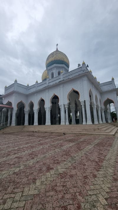 An exterior, low-angle shot shows a mosque in Kambam, Indonesia. The mosque is predominantly white with arched walkways supported by columns and topped with gold domes. The ground in front of the mosque is paved with interlocking red and grey bricks. The sky is overcast and grey. There are no people visible in the image.