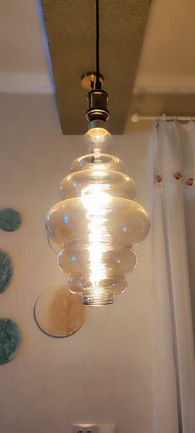 This is a medium shot of a decorative glass pendant light fixture. The light source is a visible filament bulb emitting a warm, bright glow that reflects off the tiered, amber-colored glass shade. The fixture is suspended from the ceiling by a black cord and a brass-colored holder. Behind the light, to the right, is a white sheer curtain with decorative embroidery of peaches. To the left of the light, on a light-colored wall, are three circular decorative wall hangings. The top one is a soft green with a leaf-like pattern, the middle one is a lighter color with an imprint resembling a leaf, and the bottom one is partially visible and appears to be similar to the middle one. The ceiling has a textured, light brown panel above the light fixture. The overall scene suggests an indoor setting, likely a living space or dining area, with a focus on the aesthetic of the lighting. The lighting is warm and inviting, suggesting it could be evening or a dimly lit interior. No text is visible in the image.