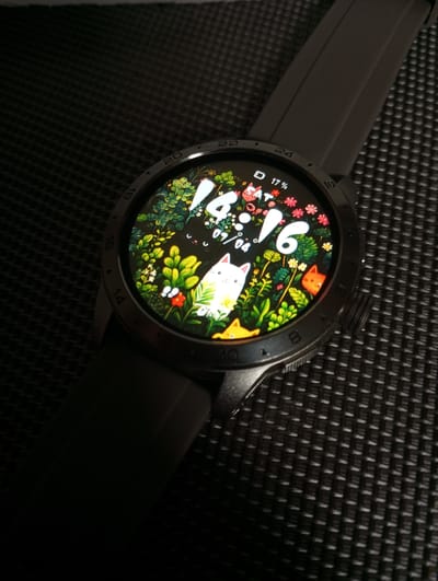 A smart watch with a whimsical digital face is resting on a textured dark surface. The watch face displays a time of 16:16, a date of 07/04, a battery indicator at 17%, and is decorated with a vibrant illustration of a lush green forest scene populated by cartoon animals. A white cat is prominently featured in the foreground, with a small orange cat peeking out from the right. The surrounding bezel of the watch shows numerical hour markers. The background surface appears to be a dark, woven material, possibly fabric or a mat, with a distinct grid pattern. The lighting suggests an indoor setting, with shadows casting across the scene. The image is a close-up, focusing on the smart watch and its display. There is no indication of the city of Dymer, Ukraine.