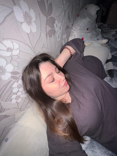 A young woman with long, dark brown hair is lying down with her eyes closed, appearing to be asleep. She is wearing a dark colored shirt or robe and a beaded bracelet on her left wrist. She is propped up by pillows, one of which is light beige and has a patterned wallpaper with floral designs behind her. To her right, there are stuffed animals and blankets, one of which is white with red heart-shaped patterns. The lighting is dim, suggesting it is nighttime or a dimly lit room. The scene appears to be indoors, possibly in a bedroom in Shirkovo, Russia.