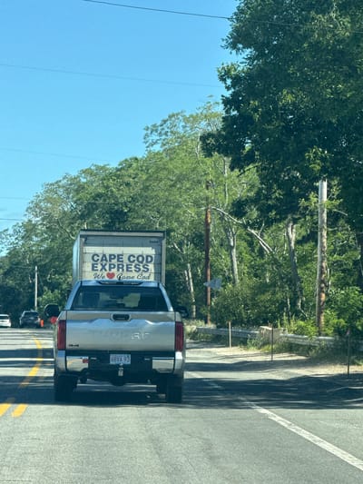The image shows a silver Toyota Tundra pickup truck with a white cargo box attached, driving on a road in Wellfleet, Massachusetts. The cargo box has "CAPE COD EXPRESS" printed on it in large blue letters, with a red heart symbol and "We Cape Cod" below. The truck's license plate reads "6BVA 93". The scene is set on a sunny day with lush green trees lining the road. Other vehicles are visible in the background, suggesting a moderate traffic flow. The road appears to be a two-lane highway with yellow lane markings.
