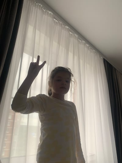 In a dimly lit room with sheer white curtains, a young girl is captured from an unusual, upside-down perspective. She is wearing a light-colored, long-sleeved shirt with a subtle pattern. Her hair is light brown and appears slightly tousled. The girl is making a peace sign with her right hand, her fingers extended in the classic V-shape. Her face is turned towards the camera, and she has a neutral expression.

The background is dominated by the translucent white curtains, which are layered and create a soft, diffused light. Behind the curtains, hints of an outdoor scene can be seen, possibly buildings, suggesting an urban or suburban environment. The overall lighting suggests it might be daytime, as the light filtering through the curtains is bright, but the interior of the room is not fully illuminated. The room appears to be a bedroom or a living space, given the presence of curtains. The image's orientation and the girl's pose suggest a playful or candid moment captured by someone positioned above her or by the girl herself with the camera held at an unusual angle.