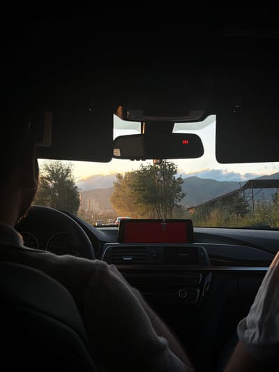 The image shows the interior of a car, with the view looking out the windshield and through the side windows. The sun appears to be setting, casting a warm, golden light on the landscape outside. Mountains can be seen in the distance, along with trees and some man-made structures. The foreground is dominated by the car's interior elements, including the dashboard, steering wheel, and a rearview mirror.
The rearview mirror displays the text "NW" in red, likely indicating a compass direction. A red object, possibly a phone or navigation device, is mounted on the dashboard. The image is taken from the perspective of someone sitting in the back seat, with a person's arm visible in the lower right corner, wearing a light-colored sleeve. Another person's head and shoulder, wearing a dark hooded garment, are also visible in the lower right and center right.
The overall impression is one of a journey during the golden hour, with a scenic view of the Armenian landscape outside. The lighting suggests it is late afternoon or early evening. The atmosphere appears calm and perhaps reflective, given the soft light and the focus on the passing scenery.