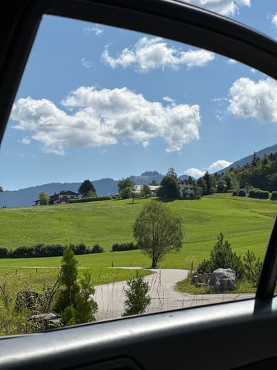 A bright, sunny day unfolds over the rolling green hills of Bischofswiesen, Germany, as viewed through a car window. The landscape is dominated by lush, vibrant green fields that undulate gently towards distant, tree-covered mountains. Several houses with traditional Bavarian architecture are nestled on the slopes, their roofs a mix of brown and red tiles. Tall, mature trees dot the fields, with one prominent tree with a wide canopy standing near the center of the frame. A winding asphalt road cuts through the scene, bordered by short, manicured hedges and clusters of smaller evergreen trees. In the foreground, the dark gray interior of a car door frames the idyllic scenery. The sky above is a brilliant blue, adorned with fluffy white cumulus clouds. The scene appears tranquil and pastoral, with no people or animals visible.