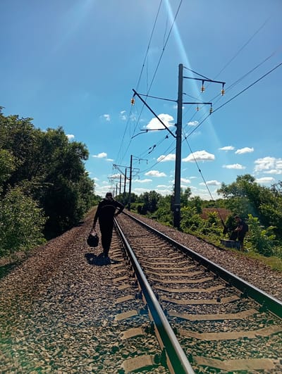 In Kolomak, Ukraine, on a bright, sunny day, two individuals are observed near a railway track. The foreground features a set of train tracks stretching into the distance, surrounded by gravel and ballast. To the left, dense green foliage lines the tracks.
In the middle ground, a person walks away from the camera along the railway track. They are silhouetted against the sky and are wearing dark clothing, carrying a black bag in their right hand. Their left hand is on their hip.
Further down the tracks, to the right, another individual is visible. They appear to be wearing dark shorts and a t-shirt, and are standing near some green bushes. They are looking down and seem to be engaged in an activity, possibly fishing, as a fishing rod is propped nearby.
Overhead, a clear blue sky is punctuated by scattered white clouds. Overhead power lines supported by utility poles stretch across the frame, indicating an electrified railway line. The sun is high in the sky, casting distinct shadows. The overall scene depicts a rural or semi-rural setting in Kolomak, Ukraine, with two people interacting with the environment near the train tracks.