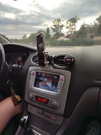 The image shows the interior of a car's dashboard, with a smartphone mounted on a stand. The car is a Ford, indicated by the design of the dashboard and infotainment system. The radio display shows "100.60 MHz" and a list of other radio frequencies. A key with a Minions keychain is visible hanging from the ignition. The car's gear shift lever is also in view. Outside the car, through the windshield, trees and houses can be seen, suggesting a rural or suburban setting. The sky is overcast with some sunlight breaking through. The context provided indicates this scene is from Shcherbani, Ukraine. The image captures a moment inside a vehicle during a drive, with the focus on the car's interior technology and its connection to the outside world.