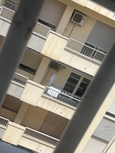 The image presents an oblique, downward-looking view of the facades of a multi-story residential building located in Ontinyent, Spain. The foreground is partially obscured by several blurred, light grey, diagonal bars, likely part of a railing or structural element.

The building exteriors are predominantly light cream or beige, exhibiting areas of weathering, staining, and discoloration. Multiple balconies with white, horizontally railed balustrades are visible across different levels. Windows are fitted with external roller shutters (persianas); some are fully closed, others are partially or entirely open.

On an upper level, a white Fujitsu brand air conditioning condenser unit is mounted externally on a wall, with black electrical conduit extending downwards. Directly below it, a balcony is present. Further down, another balcony features a white clothes drying rack with white laundry items draped over it. Adjacent to this, a single white garment, resembling a robe or tunic, hangs on a hanger against the building wall. A window on a higher level shows a small arrangement of green plants or flowers on its sill.

The scene is brightly lit by direct sunlight, indicating daytime, with strong shadows cast by architectural features. No individuals are discernible within the frame. The overall environment suggests a residential urban or suburban setting.