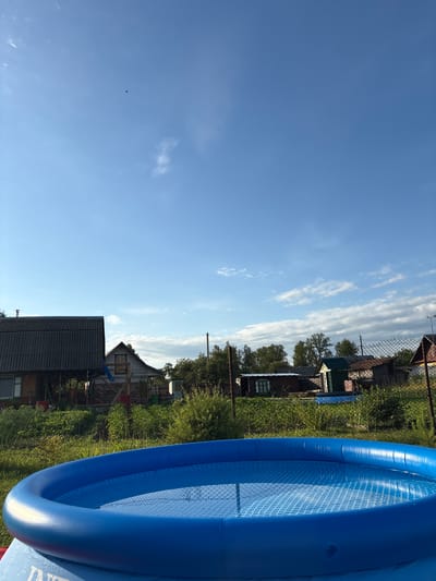 The image depicts a backyard scene with an inflatable blue pool in the foreground. The pool is partially filled with water, and its mesh lining is visible. Behind the pool, there is lush green vegetation, including tall grass and bushes. Several small structures, possibly sheds or outbuildings, are visible in the middle ground, along with a fence. In the background, there are more substantial buildings, including a house with a dark, corrugated roof and another house with a light-colored facade and a gabled roof. Trees line the horizon, and the sky above is a clear blue with scattered white clouds. A single bird is visible in flight against the sky. The scene suggests a rural or suburban setting, consistent with the location of Неманіцкі сельскі Савет, Belarus, which is characterized by its agricultural landscapes and residential areas with individual houses and gardens. There are no people or animals clearly visible in the image, apart from the distant bird. The overall atmosphere is serene and depicts a sunny day.
