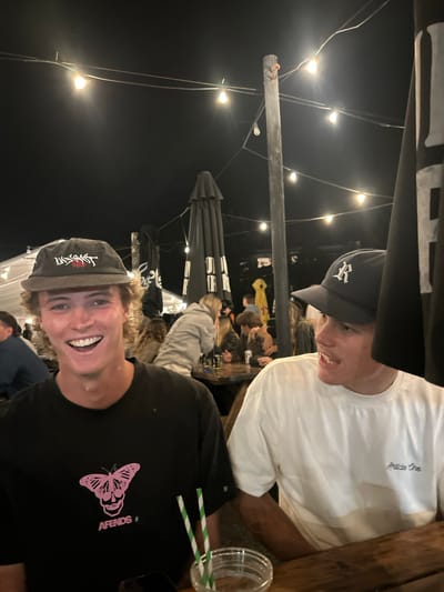 A nighttime scene depicts an outdoor or semi-outdoor establishment in Wombarra, Australia, illuminated by suspended string lights. In the foreground, two young adult males are seated at a wooden table. The male on the left, with light brown curly hair, smiles broadly towards the camera, revealing teeth. He wears a dark flat-brimmed cap with a white and red design and a black t-shirt featuring a pink butterfly-skull graphic and the text "AFENDS". A clear glass containing a drink with two green and white striped straws, and a dark smartphone, are on the table in front of him. The male on the right wears a dark blue baseball cap with a white "R" logo and a white t-shirt with "Article One" embroidered on the left chest. He is positioned looking to his left, with his right forearm visible on the table. The background shows multiple indistinct figures seated at tables, dark closed parasols or umbrellas, and wooden poles supporting the string lighting.