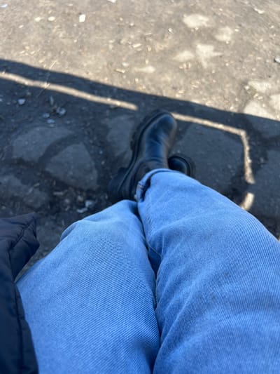 A first-person, downward-angle photograph captures the lower body of an individual seated or reclined outdoors in Большевик, Russia. The subject's legs are extended forward, clad in light blue denim jeans. Both feet wear black, chunky-soled boots. A dark, possibly black, portion of a puffy jacket or coat is visible in the lower-left. The ground surface is a rough, uneven mixture of light gray dirt or weathered pavement, containing scattered small debris. Strong sunlight casts a prominent, dark, angular shadow, consistent with a railing or similar linear structure, across the ground from the upper-left to the lower-right.