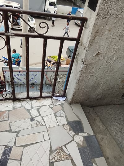 The image captures an overhead view looking down onto a bustling street scene, framed by a metal railing and a concrete wall in the immediate foreground.
In the foreground, a floor is visible, constructed from a mosaic of irregularly shaped and sized ceramic or stone tiles in shades of white, beige, light gray, and dark gray, with some tiles featuring geometric patterns. The grout lines are distinct, and some tiles appear chipped. A dark, rusty brown metal railing with vertical bars and decorative twisted scrollwork on the left runs horizontally across the upper part of the foreground, separating the viewer's immediate space from the street below. To the right of the railing is a rough, textured concrete wall or pillar, showing signs of wear and flaking. A small piece of crumpled paper, possibly white with blue lines, lies on the tiled floor near the base of the railing and the concrete wall.
Beyond the railing, at street level, is a light-colored paved road or asphalt. Several people are visible:
* A man in a light grey t-shirt and dark pants walks towards the right in the middle of the street.
* A woman in a vibrant blue patterned top and dark bottom stands on the left side of the street, near a temporary setup.
* Further back, near a white truck, two men are standing, one in a white shirt, the other in a blue shirt.
* A person is stooped or sitting near a fruit stall on the right.
Various vehicles are present: a white van is parked on the left, a white cargo truck is further back, a dark green or blue car is partially visible on the right, and another white car is in the distant background.
A market-like atmosphere is suggested by vendor stalls. On the right, a stall displays a large quantity of bright green fruits, possibly apples or limes, in crates or on a table. Below the railing, there is a structure covered with corrugated metal sheets, on which the text "DE-UNICOBOND" is clearly visible, appearing multiple times and some instances in reverse. The ground beyond the immediate street level appears unpaved and somewhat uneven, with debris.
The scene takes place during the daytime under clear, natural light, casting visible shadows. The overall impression is that of an active urban street or market area, with many elements showing signs of age and practical use.