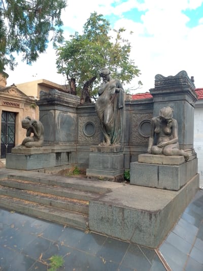 The image depicts a family mausoleum in the Recoleta Cemetery in Asunción, Paraguay. The mausoleum features three bronze statues of female figures. On the left, a seated figure is bent over with her head in her hands. In the center, a standing figure is draped in fabric and looks down. On the right, a seated figure is hunched over with her head bowed. The statues are positioned on gray stone platforms with steps leading up to them. The mausoleum structure behind the statues is gray stone with decorative circular panels. The name "VIDOVICH" is visible on a tomb in the background to the left. A tree is visible behind the mausoleum. The sky is partly cloudy. The mausoleum is situated in a graveyard with gray tiled ground in the foreground.