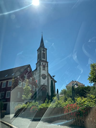 The image captures a view of the St. Lorenz Church in Schönwald im Schwarzwald, Germany. The church, with its prominent bell tower topped with a cross, is the central subject, flanked by adjacent buildings. To the left, a multi-story building with a red facade and white-framed windows is visible. Green umbrellas and lush vegetation, including red flowers, line the foreground, separated by a wooden fence. The scene is illuminated by bright sunlight, causing lens flares and reflections on what appears to be a glass surface, possibly a car window. The sky is clear and blue. The overall impression is a sunny day in a German town.