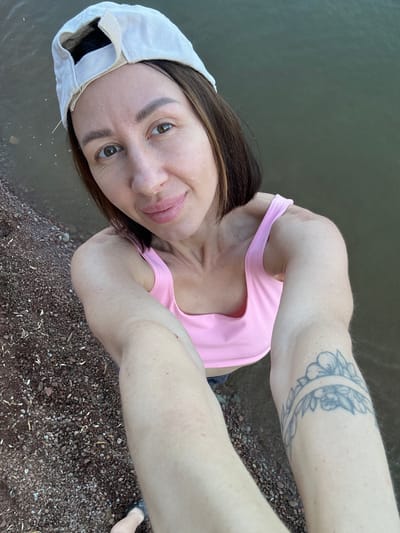 A woman wearing a white baseball cap and a pink tank top is taking a selfie. She is standing on a rocky shore with water behind her. She has a tattoo on her left arm. The location is identified as Kostovaty, Russia, which is a village in the Ryazan Oblast.
