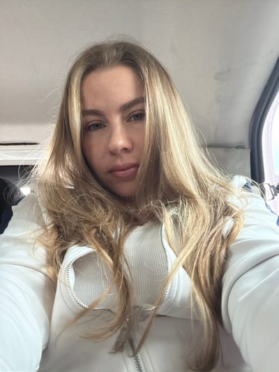 A young woman with blonde hair is pictured in a selfie. She is wearing a white zipped jacket over a ribbed white top. Her hair is long and falls around her face and shoulders. She has blue eyes and is looking directly at the camera. The background appears to be the interior of a vehicle, possibly a car or van, with a grey headliner visible above her. There is a dark rubber trim on the right side of the frame, suggesting a window or door frame. The lighting is bright, indicating it is daytime. The image does not provide any specific indicators of the location being Nova Bila, Bosnia and Herzegovina.