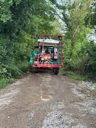 An agricultural machine, possibly a sprayer or planter, is captured on a muddy and gravelly track, surrounded by lush green foliage. The scene is set outdoors in a rural environment, consistent with Cross Keys, Ireland.
The primary subject is a large, red-framed farm implement, attached to what appears to be a tractor, though only the cab and a section of its white canopy are clearly visible. The machine features numerous green, cone-shaped fixtures arranged in rows, extending outwards from the main body. Inside the tractor's cab, a person can be faintly seen operating the vehicle. Headlights or warning lights on the front of the machine are illuminated, suggesting it is either in operation or being prepared for use. A small, yellow rectangular sign with partially illegible text (possibly "GUL") is attached to the side of the red frame.
The track itself is a mix of light-colored gravel and dark, wet mud, showing distinct tire tracks, indicating recent passage. Several puddles are present, reflecting the sky and suggesting recent rain or damp conditions. On either side of the track, dense green trees and bushes form a natural barrier, creating a verdant tunnel effect. The foliage appears thick and healthy, with some bare branches visible higher up on the left. Small white flowers can be seen growing amidst the green vegetation on the left side of the track.
The time of day appears to be daytime, with soft, diffused lighting suggesting an overcast sky or early morning/late afternoon. The overall impression is one of rural activity, with the farm machine navigating a damp, unpaved lane.