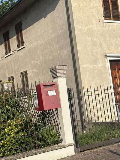 The image captures a sunlit exterior scene in Rudiano, Italy. A weathered, beige building with dark-slatted windows is the primary structure. A bright red "POSTE" mailbox stands on a concrete post to the left of a dark metal fence that encloses a patch of lush green vegetation. The fence runs horizontally across the foreground of the image. A sliver of a wooden door is visible on the right side of the building. The ground is paved with cobblestones. The lighting suggests it is daytime, with shadows cast by the building and fence.