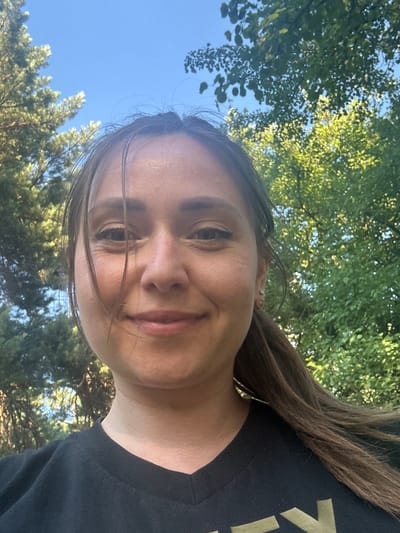 A woman is taking a selfie outdoors. She has brown hair pulled back into a ponytail and is smiling at the camera. She is wearing a black t-shirt. Behind her, there are trees and a blue sky. The scene is likely in Chmyrivka, Ukraine, given the information provided.