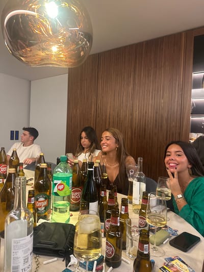 The image captures an indoor social gathering, ostensibly located within Lordelo do Ouro e Massarelos, Portugal, around a light-colored table. Four young individuals are seated. On the far left, a male with short dark hair, wearing a white t-shirt, looks towards the left side of the frame. To his right, a female with long dark hair, wearing a white top, is focused on a smartphone held in her hands. Adjacent to her, a second female with long light brown hair, in a dark V-neck top, smiles towards the right. On the far right, a third female with long dark hair, wearing a green top, looks directly at the camera with her tongue slightly extended and makes a peace sign gesture with her right hand.
The table surface is cluttered with numerous glass bottles, predominantly brown beer bottles labeled "SUPER BOCK" and "Coral Cerveja Especial," alongside a clear glass bottle labeled "TEJO," and a green 7-Up soda bottle. Multiple empty and partially filled glasses, some containing a light-colored liquid, are dispersed across the table. Also present are a black bag, several loose cigarettes, cigarette butts, and mobile phones.
The environment includes a white ceiling from which a large, illuminated, amber-tinted spherical light fixture hangs centrally in the foreground. The rear wall features vertical dark wood paneling, while the wall to the left is a plain light color. A partially visible, internally lit shelving unit is present on the far right, adjacent to the wooden paneling.