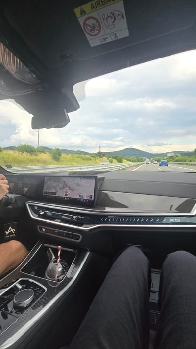 This media file captures an interior view from a car driving on a highway in Studena, Bulgaria. The perspective is from the passenger seat, looking forward. The dashboard is dominated by a large infotainment screen displaying a map, and to the right, a textured carbon fiber panel with illuminated blue accents and the "X5" logo. A glass with a red and white striped straw rests in a cup holder. In the foreground, a person's legs, clad in dark jeans, are visible.
Outside the car, the scenery reveals a highway with other vehicles. The road is flanked by grassy fields and rolling hills covered in lush green trees. The sky is partly cloudy with a mix of bright sunlight and diffused light, suggesting a pleasant, possibly late afternoon, weather. The overall atmosphere appears calm and ordinary, indicative of a typical road trip. An "AIRBAG" warning sticker is visible on the sun visor. The time of day appears to be daytime, with good visibility.