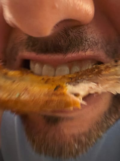 A close-up image shows a person eating. The person's mouth is open, revealing teeth and facial hair. A piece of food, possibly meat on a bone, is being held and consumed. The texture and color of the food are visible. Part of a hand is also visible, seemingly holding the food. The background is blurred.