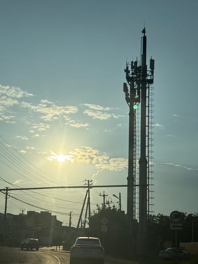 The image depicts an outdoor scene with a tall industrial structure, possibly a telecommunications tower or a crane, dominating the sky. The structure is silhouetted against a bright, hazy sky, suggesting it's either early morning or late afternoon. The sun is visible, low in the sky and partially obscured by clouds, casting a warm glow. Scattered clouds are present, some appearing wispy and others more substantial, illuminated by the sunlight.
Below the structure, there are visible power lines and poles, indicating the presence of infrastructure. In the foreground, a road is visible with a few vehicles, one of which is a car with its brake lights illuminated. The road appears to be wet, suggesting recent rain or a damp surface. The overall setting seems to be semi-urban or industrial, with some hints of buildings or structures in the background.
A traffic sign is visible on the right side of the image, showing a truck symbol with a diagonal line through it, indicating a restriction for trucks. Another sign with an arrow is also visible. The time of day is likely late afternoon due to the position of the sun and the angle of the light. The weather appears to be clear with some cloud cover. The scene conveys a sense of everyday transit or industrial activity under a soft, diffused light. There are no people explicitly visible in the frame.