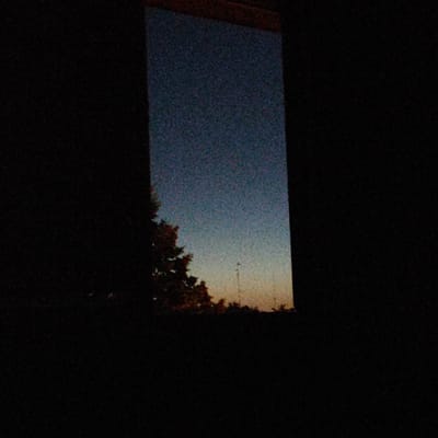 The image depicts a view through a window at dusk. The sky outside is a gradient of deep blue at the top, transitioning to a lighter blue and then a warm orange-yellow near the horizon. The sky is grainy, suggesting low light conditions or a photographic artifact. On the left side of the frame, a dark silhouette of a tree with foliage is visible against the sky. In the distance, faint outlines of what appear to be buildings or structures are present, along with a vertical element that could be a lamppost or utility pole. The foreground and immediate surroundings of the window are completely dark, obscuring any details within the room or immediate exterior. The overall mood is somber and still, with the fading light indicating the end of the day. The location context provided is Płock, Poland. There is no visible text in the image.