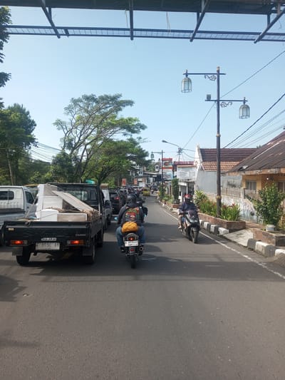 The image shows a street scene with vehicles and pedestrians in what appears to be a busy area. The location is likely in Indonesia based on the license plate, it is not in Khe Luang, Laos. A black Suzuki Carry pickup truck loaded with furniture is in the foreground, followed by other cars and motorcycles. Two motorcycles are prominently visible, one directly behind the truck and another driving in the opposite direction. Buildings and trees line the street, and a decorative street lamp is on the right side of the road. The sky is clear and blue.