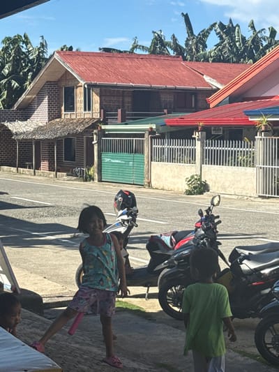The image depicts a street scene in Burgos, Philippines, under bright daylight. In the immediate foreground, a paved concrete surface shows two children and several parked motorcycles. A young girl with short dark hair, wearing a light blue patterned tank top, pink shorts, and pink flip-flops, stands smiling towards the camera with her left leg extended backward. To her right, a young boy in a green t-shirt and light-colored shorts stands mostly with his back to the camera, facing the right side of the frame. A third child's head is partially visible at the bottom left of the frame, resting on a white surface.

Three motorcycles or scooters are parked on the right side of the paved area. A white and black scooter in the center foreground has a black motorcycle helmet resting on its handlebars. To its right, a black scooter is draped with a red and blue fabric item over its seat. Strong shadows from an unseen structure are cast across the foreground pavement.

A dark gray paved road with a white center dividing line separates the foreground from a row of residential structures. Across the road, on the left, stands a two-story house with a red corrugated metal roof. Its upper story features a light-colored brick facade with windows, while the lower story has a similar brick facade and a thatched roof extension over a porch area, supported by brick columns. Adjacent to it, partially obscured by a white concrete fence, is a section of a house with a green roof, fronted by a green metal gate. To the right, another house with a red corrugated metal roof is visible, fronted by a white concrete fence topped with several potted plants. Dense tropical foliage, primarily banana trees, rises behind the houses against a clear blue sky.
