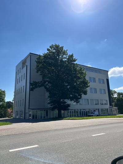 A daytime, eye-level, medium shot shows a modern, multi-story office building on a clear day in Tartu, Estonia. The building is light gray with numerous rectangular windows and a darker vertical trim. A large, mature green tree stands in front of the building, partially obscuring it. The foreground features a paved sidewalk and a gray asphalt road with white lane markings. A white van is parked in front of the building. The sky is a bright blue with scattered white clouds and the sun is visible in the upper right corner, creating lens flare. The surrounding area appears to be a developed urban or suburban environment with other buildings and trees visible in the distance.