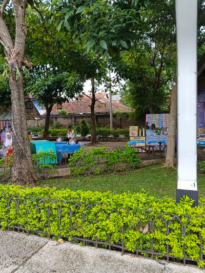 This image depicts an outdoor public area, identified as being in Bien Hoa, Vietnam, characterized by a mix of natural elements and informal commercial activity. In the foreground, a textured gray paved surface is visible on the bottom left, bordered by a low black wrought iron fence obscured by dense bright green foliage. A prominent tree with a thick brown trunk and epiphytic plants on its bark stands on the left.
The midground features a grassy area interspersed with green bushes. Two large, irregularly shaped blue concrete blocks, possibly seating or decorative, are centrally located. To the left, a female individual wearing a white hijab and light-colored clothing is seated on a low stool or bench. Behind the blue blocks, a male individual in a white shirt and dark pants is seated on a low stool, positioned as if attending to a small stall. This stall includes items and signage, with visible text resembling "Pudini Kurma" and "Kurma". Further to the right, a bright white rectangular pillar or lamp post stands erect. Several additional vendor stalls or tables are visible behind the grassy area on the right, displaying an assortment of packaged goods, bottles, and various products.
In the background, a concrete wall separates the public area from residential structures, identifiable by their reddish-brown tiled roofs. Numerous trees with green foliage are scattered throughout the scene, and the sky appears overcast. The scene suggests a relaxed public space with small-scale vending operations.
