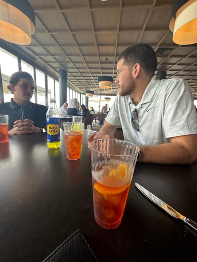 Two men are at a restaurant table. One wears a white shirt and sunglasses, while the other wears a black shirt. A pitcher of orange drink sits on the table with a filled glass and a bottle of Inca Kola. A knife and a black placemat are also visible. The restaurant has a high ceiling with rectangular panels and modern hanging lamps. Other patrons can be seen in the background, seated at various tables.