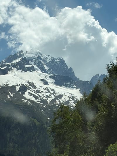 The media file shows a dramatic mountain landscape. The primary subject is a large, snow-capped mountain with rocky outcrops and patches of snow. The mountain dominates the right and central portions of the image, with a steep, rugged face. On the left, fluffy white clouds are set against a bright blue sky. The top right of the frame is partially obscured by dark green foliage, likely trees or bushes.

The scene suggests an outdoor activity, potentially hiking or mountaineering, as there appear to be faint tracks or paths visible on the snow-covered slopes of the mountain. There are also small, dark figures visible on the mountain, which could be people engaged in these activities. The overall impression is one of a grand, natural setting.

Notable details include the contrast between the bright blue sky and white clouds with the darker, shadowed mountain. The sunlight appears to be strong, casting shadows on the mountain face. The time of day is likely daytime, given the brightness of the sky. The presence of snow indicates a high altitude or a cool climate. The foliage suggests that the lower slopes of the mountain are forested.

There is no visible text in the image. The location context of Vallorcine, France, suggests this is likely a view of the French Alps.