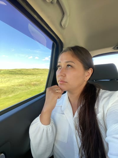 A young woman with long, dark hair pulled back into a ponytail sits in the passenger seat of a vehicle, looking out the window. She is wearing a white collared shirt with puffed sleeves and a ring on her right hand. The window shows a blur of green fields under a bright blue sky with scattered white clouds. The interior of the car is visible, including a dark headrest behind her and a portion of the ceiling. The scene appears to be a snapshot of a road trip through a rural area, possibly near the village of Tazlarovo, Russia, known for its natural landscapes.