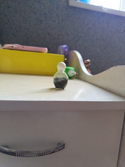A small, clear glass bottle with a white cap sits on a light-colored surface, filled with dark granules. Behind it, a bright yellow rectangular object, possibly a case or box, is visible, with a pink object, resembling a phone, resting on top. Further back, a green tube and a blurry, small figurine are present. The background consists of a textured blue wall and a window with light streaming through. The foreground shows the edge of a white drawer with a decorative handle made of shiny, patterned metal. The scene appears to be indoors, possibly on a dresser or shelf, and the overall impression is one of domestic clutter. The image does not provide any specific details that would allow for its location to be identified in the city of Shirin, Uzbekistan.