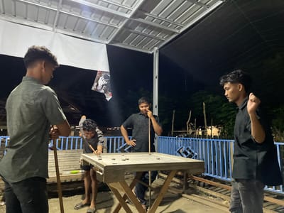 Four young men play a game of disk-pool outside at night. The men are standing and holding pool stick style cues around a raised wooden table with disks on top. In the background are blue metal railings and a dense treeline. The location is the city of Khang Tanjong, Indonesia.