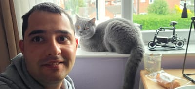 A male individual is captured in a close-up selfie in an indoor setting within the city of Oswestry, United Kingdom. He is positioned on the left side of the frame, facing slightly right towards the camera, and exhibits a subtle smile. He wears a grey hooded sweatshirt. Behind and to the right of the man, a grey British Shorthair cat with amber eyes is seated on a white windowsill, looking to its left. Through the white-framed window, a residential area is visible with green foliage and houses under daylight. On the windowsill to the right of the cat, a black metallic sculpture depicting a figure riding a motorcycle is present. The wall below the windowsill is painted light purple. In the lower right foreground, a light-colored wooden surface (likely a table or desk) holds a clear glass containing liquid and small particles, and a pastry wrapped in clear plastic. A black metallic stand or tripod leg is partially visible on the far right. A brown curtain hangs on the left side of the window.