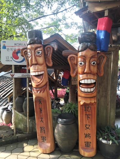 The image depicts two large, carved wooden totems resembling human figures with exaggerated facial features, specifically wide smiles and large ears, placed outdoors. Both totems are brown and have dark caps. The totem on the left has vertical black text inscribed on its lower section. The totem on the right also has vertical black text. Behind the totems, a rustic wooden structure with a pitched roof is visible, and red and blue fabric banners hang from it. To the left of the totems, a white sign displays "KOREA" and Korean text, along with a stylized image of a Korean traditional dress and a small circular emblem. Below this, another sign with an arrow points to "Thailand." Various large ceramic pots and planters containing greenery are arranged around the totems and the structure. The ground is paved with hexagonal tiles. The overall setting appears to be an outdoor tourist or cultural attraction, possibly a garden or a themed area, given the presence of decorative elements and signage. The sunlight suggests it is daytime. The location is identified as the city of Gudangkahuripan, Indonesia.