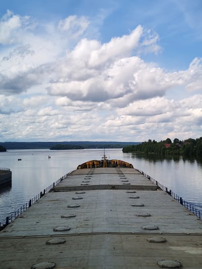 The image captures a view from the deck of a barge moving down a wide, calm river. The barge's flat, grey deck dominates the foreground, featuring a series of circular hatches. A dark railing lines the edge of the deck. In the middle ground, buoys mark the waterway. The riverbanks are lined with dense, dark green forests, with a cluster of red-roofed houses visible on the right bank. The sky above is a bright blue, dotted with numerous fluffy white clouds and some darker grey clouds, indicating a partly cloudy day. The overall impression is one of a serene journey on a river, possibly in the region of Анхимово, Russia, given the presence of such waterways and forested landscapes. There are no people visible in the image, and the barge appears to be the primary subject.