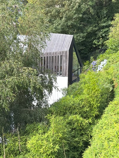 This media file depicts an outdoor scene dominated by lush green foliage and a modern architectural structure. The primary subjects are the dense vegetation, which includes trees with abundant leaves and a prominent bamboo grove, and a building with a distinctive angled roof and a facade clad in horizontal wooden slats. The building appears to be a contemporary house or annex, with large windows that reflect the surrounding greenery.
The scene is set in a natural, possibly suburban or rural, environment. The sunlight suggests it is daytime, likely a clear or partly cloudy day, casting shadows and illuminating the vibrant green of the plants. There are no people visible, nor any specific activity taking place. The focus is on the interplay between the built environment and the natural landscape.
Notable details include the geometric design of the building, the texture of the wooden cladding, and the contrast between the varied types of greenery. A cluster of small, pale purple flowers can be seen nestled within the foliage in the upper left portion of the image, adding a subtle touch of color. The overall impression is one of serenity and modern design integrated with nature.
No visible text is present in the image. The location context provided is La Clarté, France, which aligns with the aesthetic of a contemporary structure in a scenic natural setting.