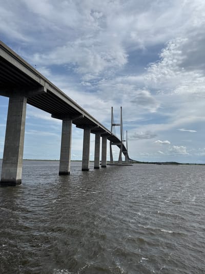 The image shows a view of the Sidney Lanier Bridge, a cable-stayed bridge that spans the Brunswick River in Brunswick, Georgia, United States of America. The bridge is constructed of concrete and steel, with tall pylons supporting the roadway. The water below is dark and choppy, with small waves visible. The sky is cloudy with patches of blue peeking through. The bridge appears to be made of concrete and is elevated over the water. There are no people or vehicles visible in the image. The scene depicts a large infrastructure project over a body of water under a partly cloudy sky.